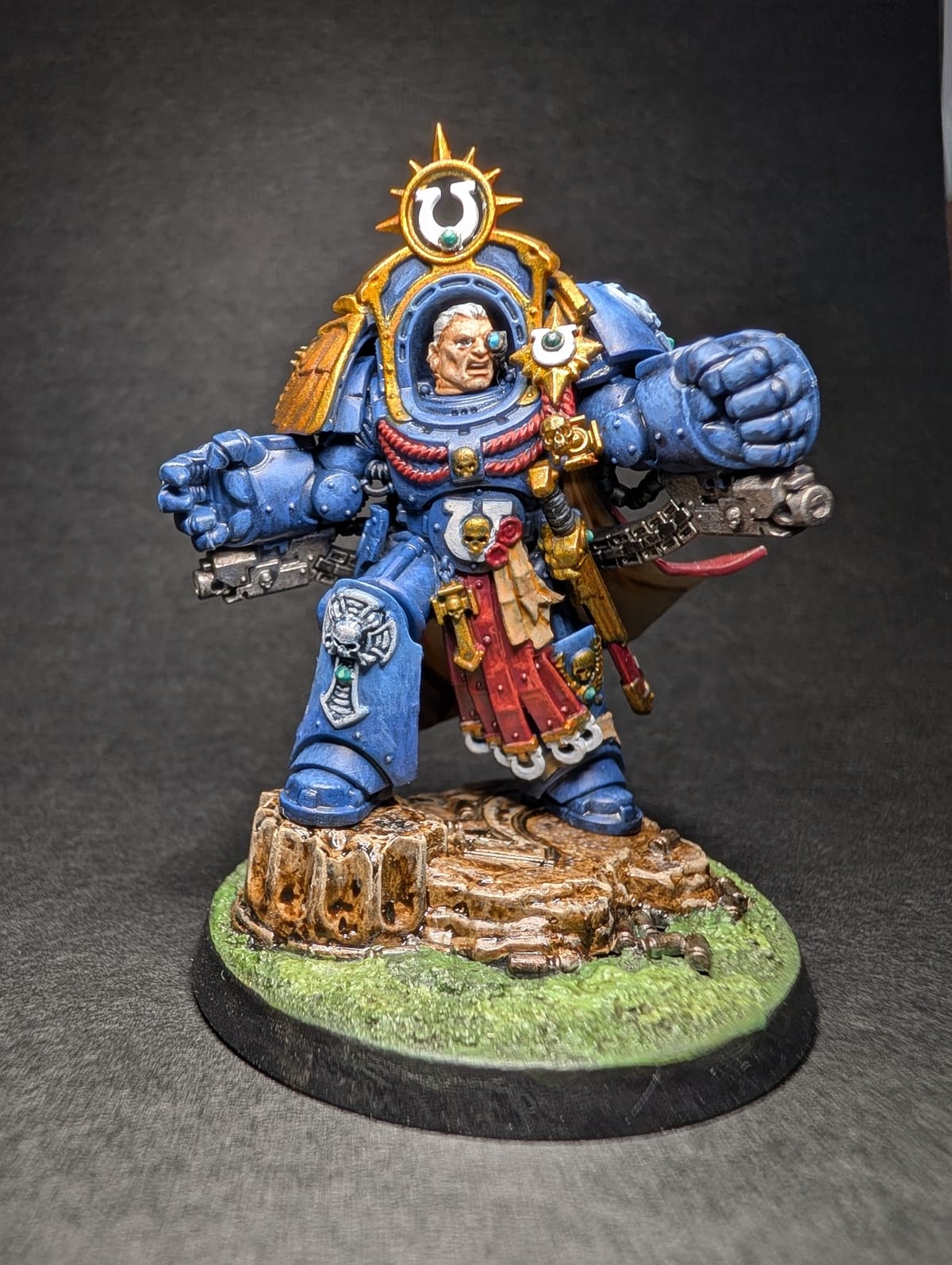 Marneus Calgar in blue Ultramarines armour with red and gold detail and twin power fists, on a muddy rocky base
