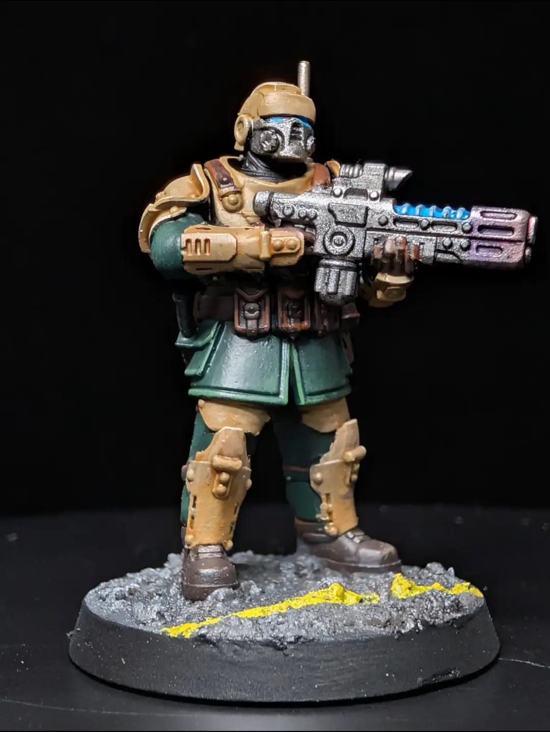 Astra Militarum Kasrkin trooper in tan and green armour with a hellgun, on a grey urban base with yellow hazard markings
